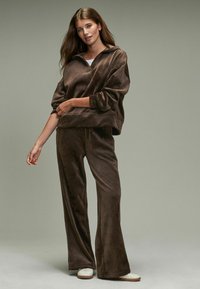 Bruin velours lounge set met een oversized capuchontrui en wijde broek, met een zachte textuur en een relaxte pasvorm. Geaccentueerd met een witte top eronder.