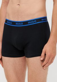 Črne bombažne bokserice z modrim elastičnim pasom, na katerem je ponovljeno označevanje "HUGO". Gladka tekstura in prilegajoča zasnova.