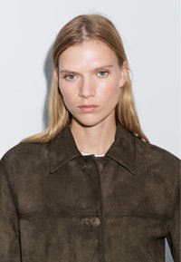 Dark brown suede jacket with a collared neckline, front button closure, and minimalistic design, featuring smooth texture and stitched accents.