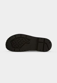 Semelle de botte noire avec des rainures profondes et un motif ondulé pour l’adhérence sur un fond clair uni.