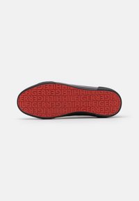 Semelle d'une chaussure noire avec une semelle en caoutchouc rouge ornée du texte en relief répété "HILFIGER" dans différentes orientations.