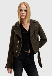 Giacca biker in suede verde oliva con zip, bottoni a pressione e tasche laterali. Abbinata a un top in maglia nera e jeans scuri.