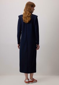 Tumes sinise värvi pikka varrukat kleit õlavarrepatjadega, sirge siluett, tagumine õmbluse detail, kantud oranžikirjadega tossudega. Sile tekstuur.