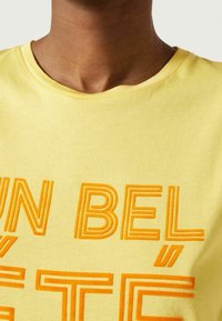 Žluté bavlněné tričko s kulatým výstřihem, které je zdobené výrazným oranžovým vyšívaným textem. Měkká textura, volný střih a mírná elasticita.