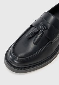 Black leather loafer with tassel accents, rounded toe, subtle stitching detail, and a textured rubber sole. Smooth finish overall.
