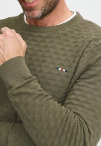 Olijfgroen sweater met gestructureerde dambordpatronen, geribbelde mouwen en een klein geborduurd logo met blauwe, witte en rode strepen.