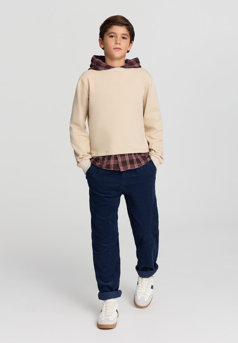 Felpa con cappuccio beige con fodera a quadri marrone scuro, indossata sopra una camicia a quadri. Abbinate a pantaloni di velluto a coste blu navy e sneaker bianche con accenti marroni.