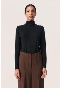 Must mustreiga pikkade varrukatega turtlenecki pood, mis on paaritud pruunide kõrge vöökohaga pükstega. Kangas näib olevat sile ja sobib kehakujusse.