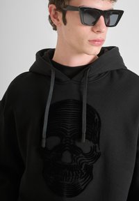 Felpa nera con un design di teschio testurizzato. Presenta un cappuccio con cordini e polsini a costine. Il modello indossa occhiali da sole neri.