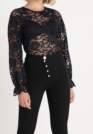 Femme portant un haut en dentelle florale noire à manches longues et un pantalon noir taille haute avec quatre boutons blancs à l'avant.