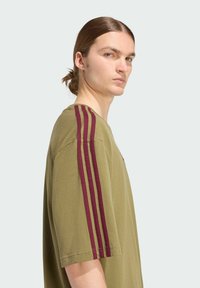 Bavlněné tričko v olivově zelené barvě s bordó svislými pruhy na ramenech, kulatý výstřih a volný střih. Hladká textura, krátké rukávy.