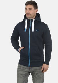 Marineblå zip-up hoodie med høj krave, kontrastblå lynlås og hvide snore. Har frontlommer og et logo på brystet.