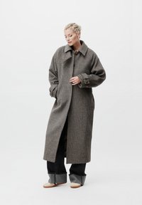 Γυναίκα που φοράει ένα μακρύ, καρώ παλτό με μεγάλα κουμπιά, φαρδιά μαύρα παντελόνια και μπεζ παπούτσια, standing against a plain white background.