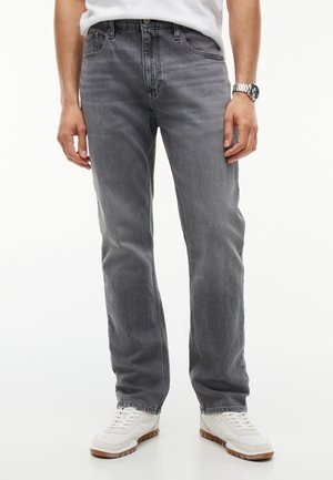 Homme portant un jean noir délavé, des baskets blanches, un t-shirt blanc et une montre-bracelet en argent, debout devant un fond blanc.