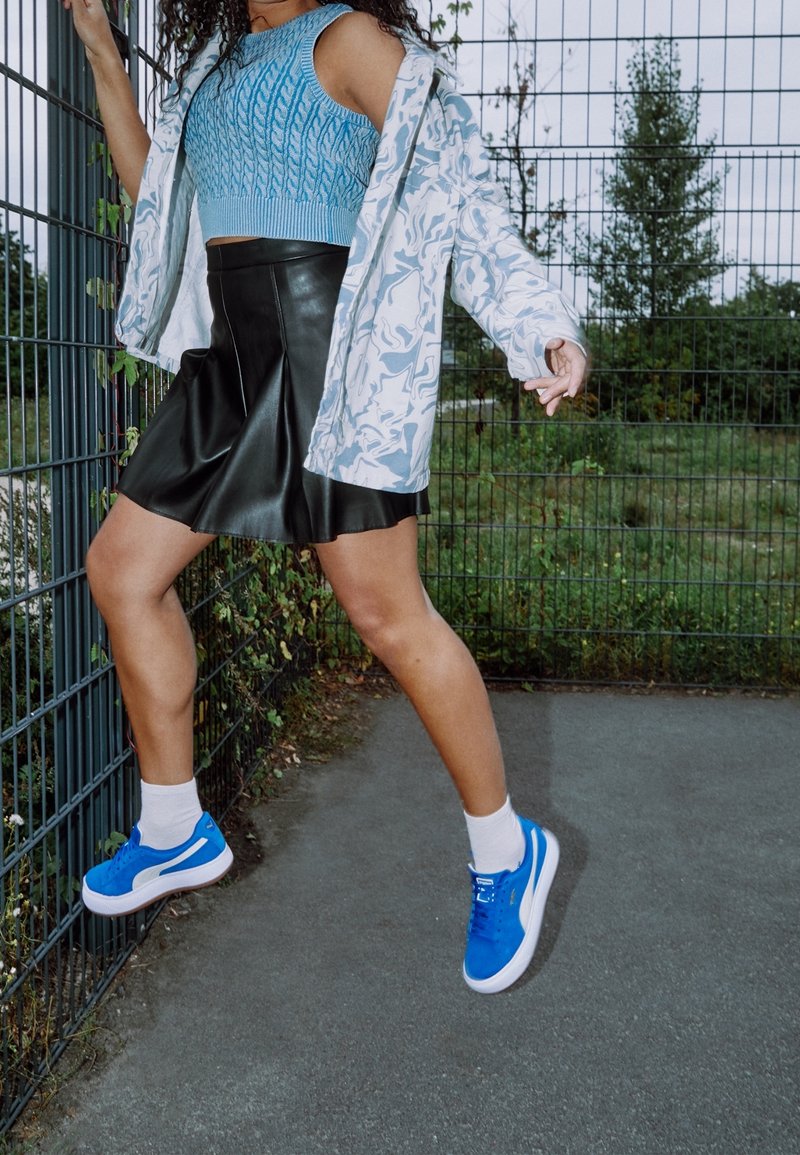 Personne portant un haut en tricot sans manches bleu, une jupe en cuir noire, une veste blanche à motifs et des baskets bleues montant sur une clôture en métal à l'extérieur.