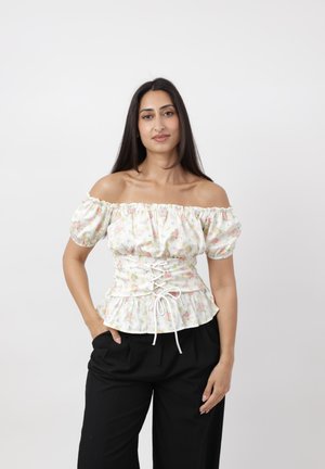 Frau mit langen dunklen Haaren, die eine weiße florale schulterfreie Bluse mit Korsett-Detail und schwarze Hosen trägt, posiert vor einem einfarbigen Hintergrund.