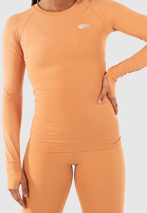 Woman wearing fitted long-sleeve peach sportswear with thumb holes, standing with one hand on hip and the other relaxed by side.