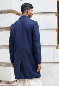 Abrigo de lana azul marino con un corte entallado, abertura sencilla en la parte posterior, solapas de muesca y textura suave, llevado sobre pantalones de color claro.