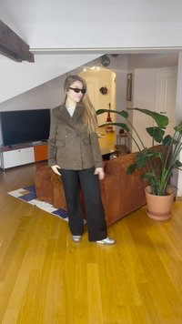 Ανοιχτό καφέ διπλόσπαθο σακάκι με δύο τσέπες μπροστά, μαύρο παντελόνι φαρδιάς γραμμής και ασημένια παπούτσια, σε ένα ζεστό εσωτερικό χώρο με φυτά.