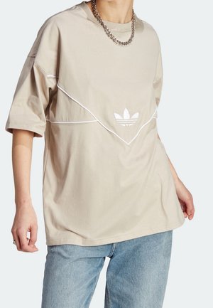 Osoba nosí béžovou oversized košili s krátkými rukávy s bílým lemováním a logem Adidas trefoil, kombinovanou s světle modrými džínami a řetízkovým náhrdelníkem.
