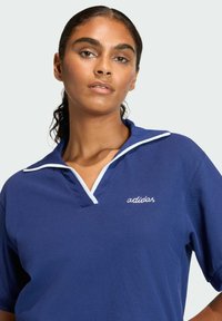 Granatowa koszulka polo z białymi wykończeniami i kołnierzykiem, z haftowanym logo "adidas" na lewej piersi. Materiał bawełniany, krótkie rękawy, luźny krój.