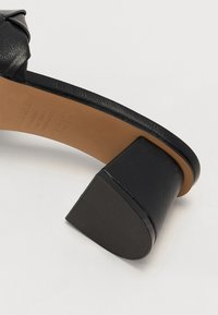 Black heeled sandal made of leather, featuring a knotted upper design, a tan insole, and a square, textured heel for stability.