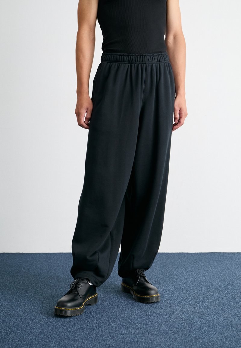 Pantalons noirs amples avec une taille élastique, fabriqués en tissu doux. Associés à des chaussures noires à lacets avec un accent jaune.