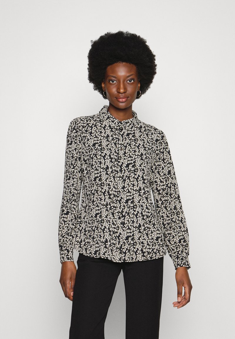 Blusa floral em preto e branco, com mangas longas, colarinho e punhos franzidos, apresentando um tecido leve e texturizado.