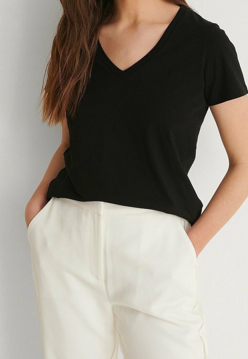 Vrouw draagt een zwart T-shirt met korte mouwen en V-hals, ingestopt in heuphoge witte broek, met handen in de zakken.