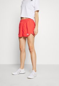 Röda sportiga shorts med elastisk midja och sidofickor, prydda med The North Face-logotypen på nedre vänstra sidan. Bärs tillsammans med en vit kortärmad T-shirt.