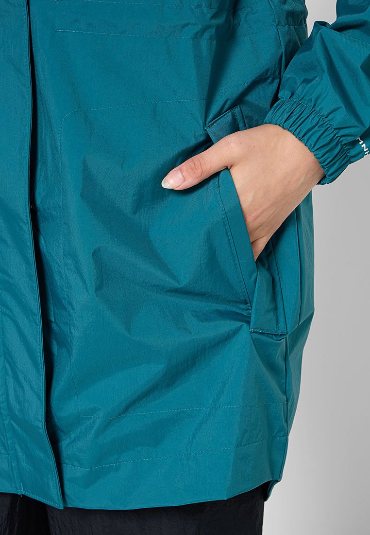 Main dans une poche d'une veste de pluie turquoise avec des poignets élastiques sur les manches et un pantalon noir visible en dessous.