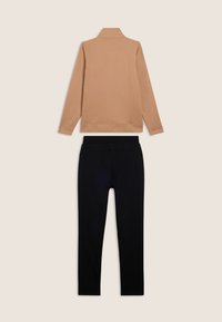 Maglietta a maniche lunghe color cammello con colletto alto abbinata a pantaloni neri con vita elastica, disposti su uno sfondo beige.