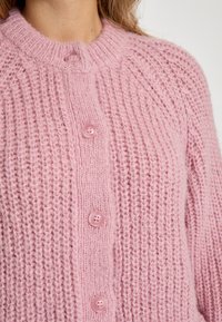 Roosa kudumine kardigan, millel on tekstuuriline muster, soonikkoes krae ja viis roosat nuppu ees. Pehme, karvane kangas ja vabalt istuv lõige.