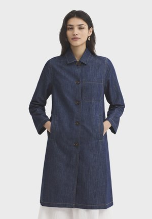Femme aux cheveux foncés portant un manteau en denim bleu foncé boutonné avec col, poches poitrine et poches pour les mains, debout devant un fond blanc uni.