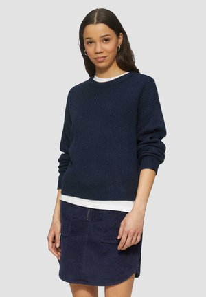 Marineblauwe gebreide trui met verlaagde schouders, gecombineerd met een donkere corduroy rok met een voorzak met rits en een gestructureerde stof.