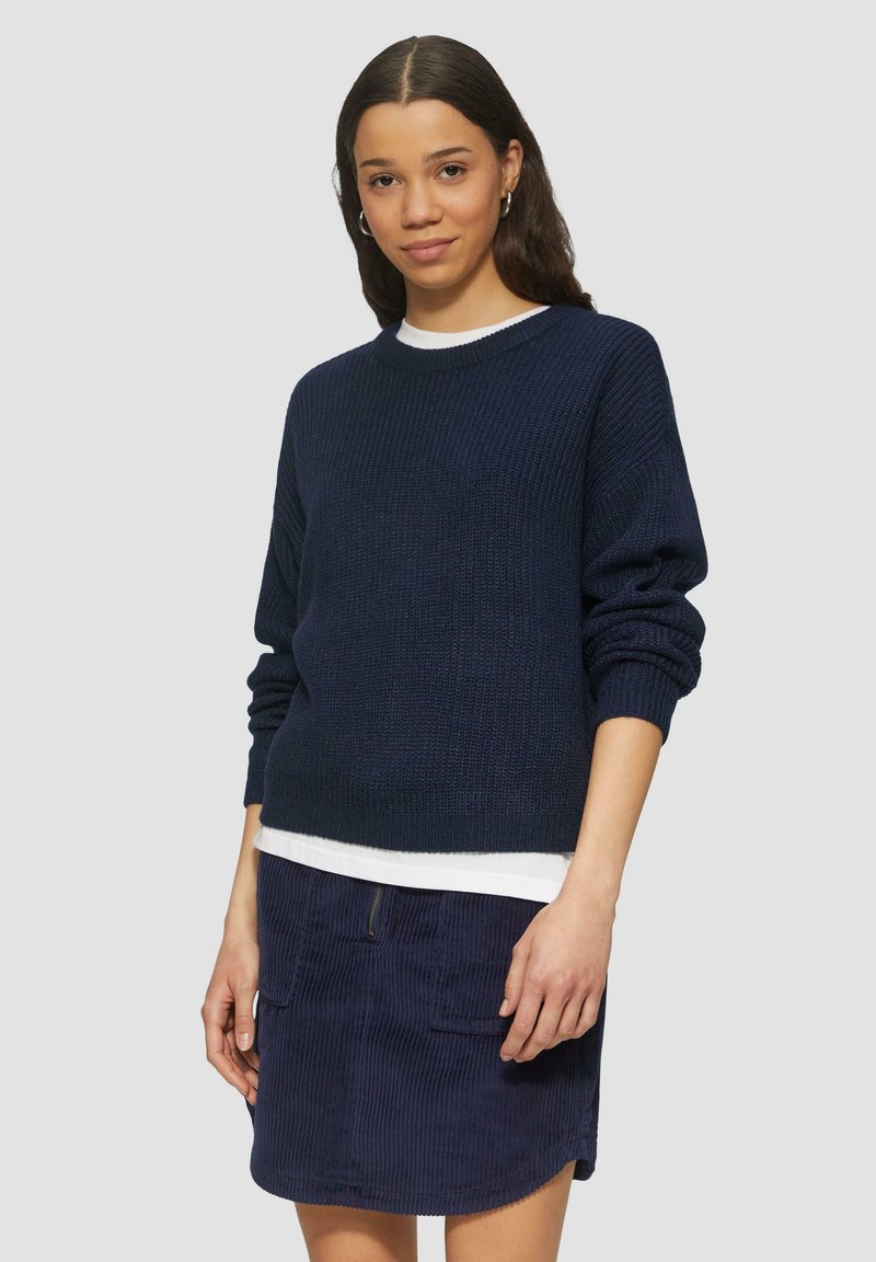 Pull en tricot bleu marine avec épaules tombantes, associé à une jupe en velours côtelé foncé avec une poche zippée devant et un tissu texturé.