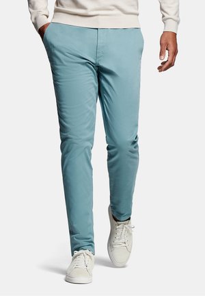 Man loopt en draagt lichtblauwe strakke broek, witte sneakers en een crème trui met de handen gedeeltelijk in de zakken.