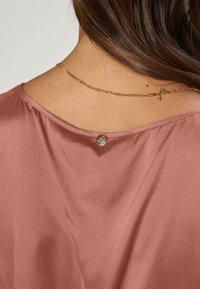 Närbild av en persons övre rygg med brunt hår, iförd en rosafärgad satinblus och ett tunt guldhalsband.