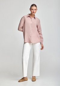 Camicia button-up rosa con vestibilità rilassata e maniche lunghe, abbinata a pantaloni bianchi a gamba larga e scarpe color cammello. Design semplice, texture morbida.