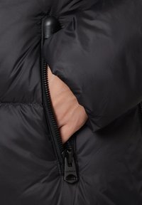Ręka włożona do częściowo rozpiętej kieszeni czarnej, ocieplanej kurtki z widocznym czarnym suwakiem.