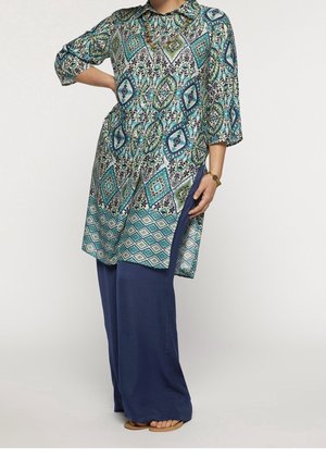 Kobieta ubrana w tunikę w geometryczne wzory w kolorach niebieskim, zielonym i białym, w połączeniu z luźnymi, ciemnoniebieskimi spodniami oraz sandałami.