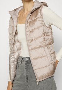 Chaleco ligero acolchado en rosa suave, con cierre de cremallera frontal, capucha y un sutil patrón interior. Diseño sin mangas con bolsillos laterales.