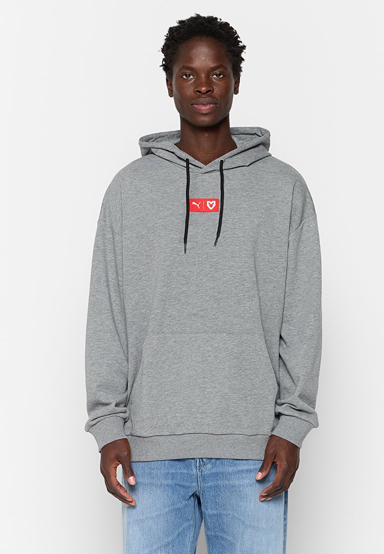 Sudadera gris de corte relajado, con un parche de logo rojo en el pecho, cordones negros y puños de canalé. Llevada con vaqueros de un tono azul claro.
