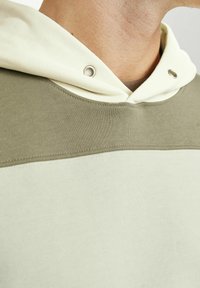 Sweat à capuche avec une capuche crème et un corps vert olive. Il présente une texture lisse, des détails côtelés et des œillets en métal sur la capuche.