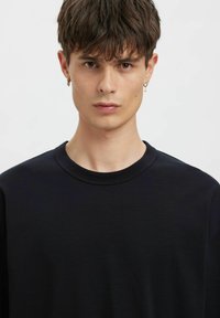 Melna adīta T-krekla ar apaļu kaklu, kam ir gluda tekstūra un īsas piedurknes. Drēbju tuvplāns uz modeļa, kurš valkā auskarus.