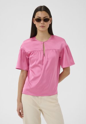 Kobieta o długich ciemnych włosach, nosząca beżowe okulary przeciwsłoneczne, jasnoróżową bluzkę z krótkim rękawem i przednim rozcięciem oraz jasnobeżowe spodnie na jednolitym tle.