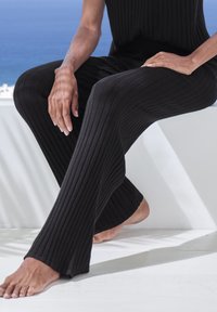 Schwarze, gerippte Strickhosen haben ein ausgestelltes Design mit einer strukturierten Oberfläche. Die nackten Füße des Models ruhen auf einer weißen Fläche vor einem blauen Hintergrund.