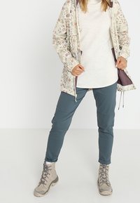 Kevätkuvioinen kevyt takki valkoisen villapaidan päällä, yhdistettynä teal-värisiin housuihin ja beigeihin retkeilykenkiin, esittelee rentoa ulkoilutilaa.