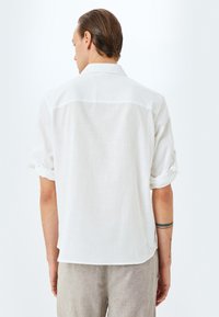 Camisa blanca de lino con mangas enrolladas, que presenta una textura suave y un sutil patrón tejido. La vista trasera muestra un dobladillo redondeado.