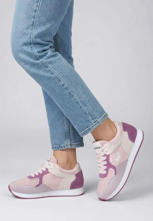 Personne portant un jean bleu clair et des baskets rose pastel-violet, debout avec un pied légèrement levé.